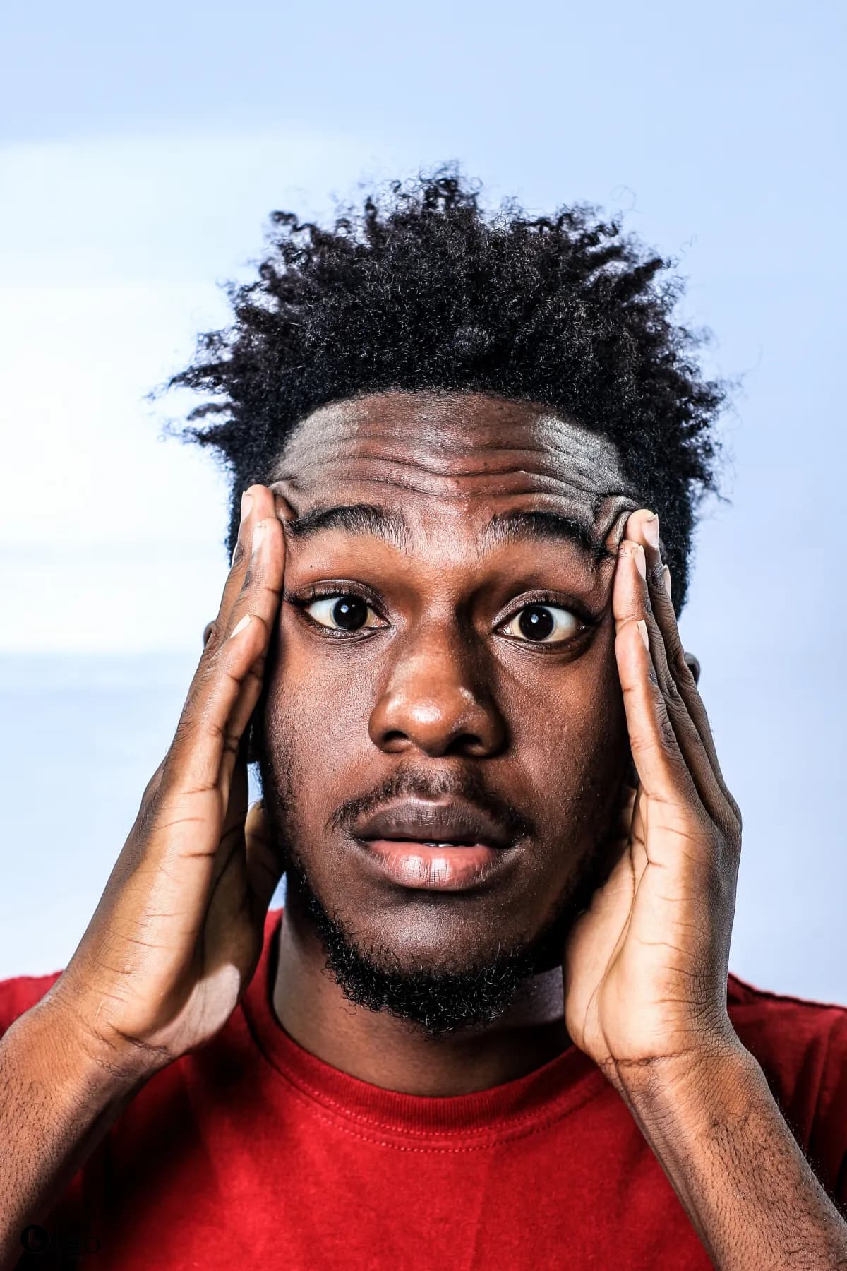 A man in a red shirt with his hands resting against his temples.